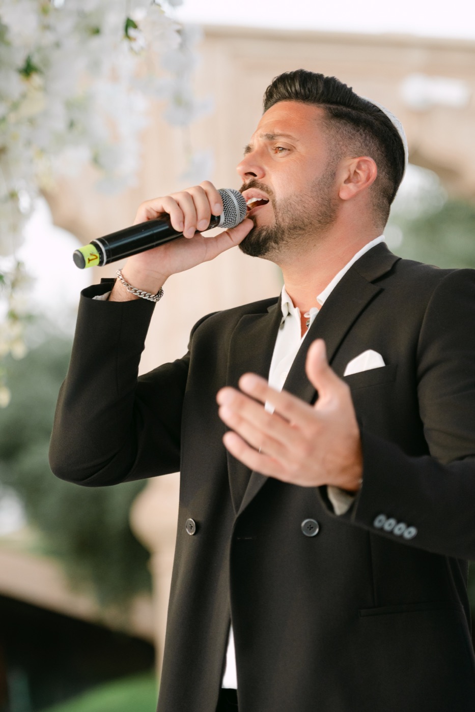 Chanteur en costume noir au micro, fleurs blanches en arrière-plan, Drop Music