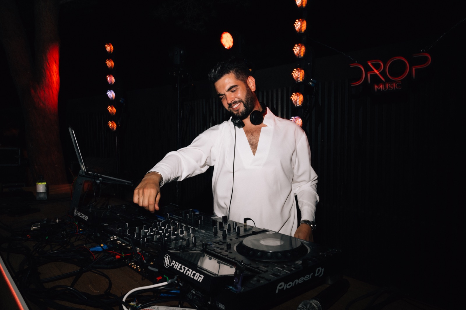 DJ en chemise blanche souriant aux platines, logo Drop Music au fond, Drop Music