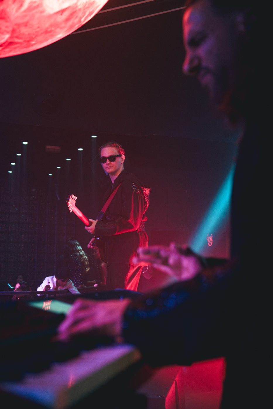 Bassiste sur scène sous une lumière rouge, clavier au premier plan, Drop Music