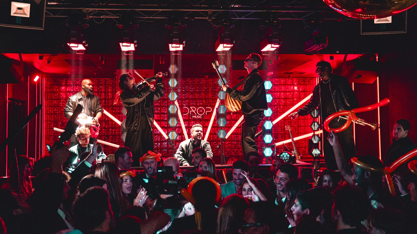 Cuivres et chanteurs sur une scène éclairée en rouge, invités au premier plan, DJ Live Drop Music