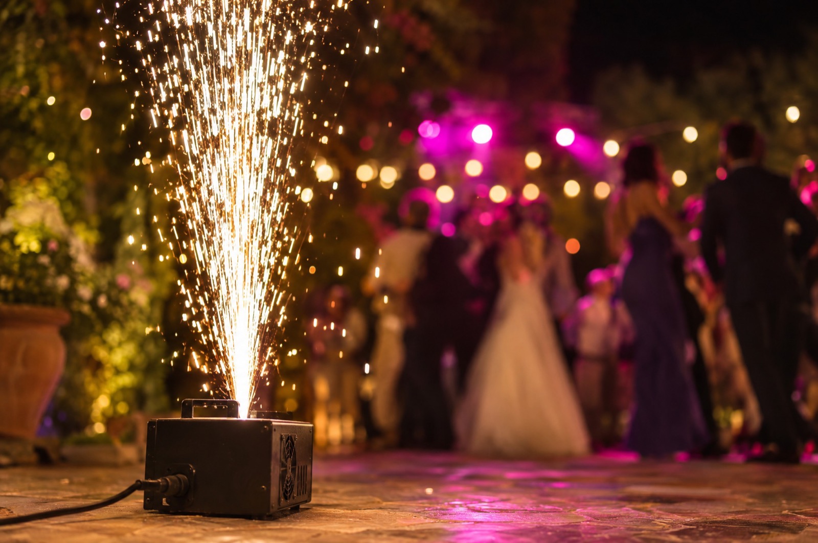 Machine à étincelles froides au premier plan dans un jardin, jet blanc vers le ciel, la mariée et les invités derrière sous les guirlandes, Drop Music